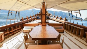 A wooden deck with a central table and benches, surrounded by a covered area with a view of the sea and mountains. - Tanaka Liveaboard (Labuan Bajo)