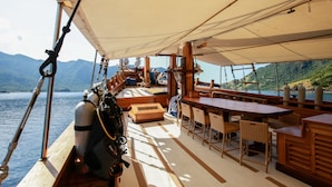A boat's deck with a dining area, seating, and a view of the water and mountains. - Tanaka Liveaboard (Labuan Bajo)