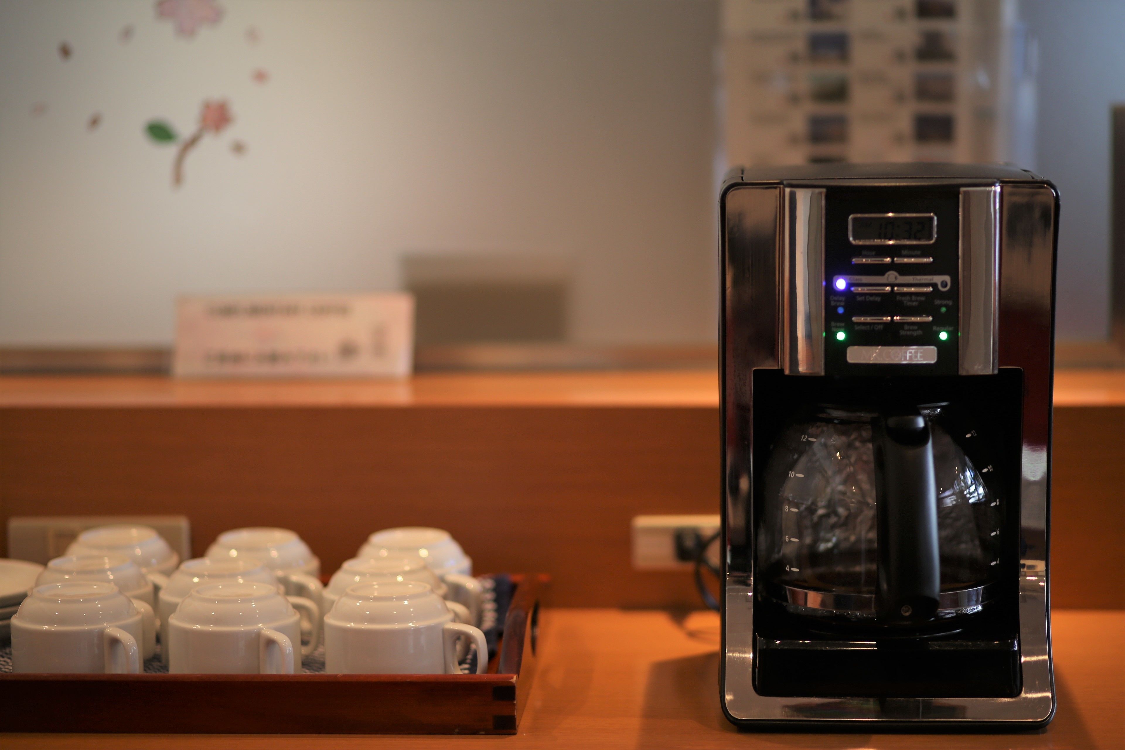 Máquina de café com visor digital e jarra, sobre bancada de madeira com bandeja de chávenas brancas.