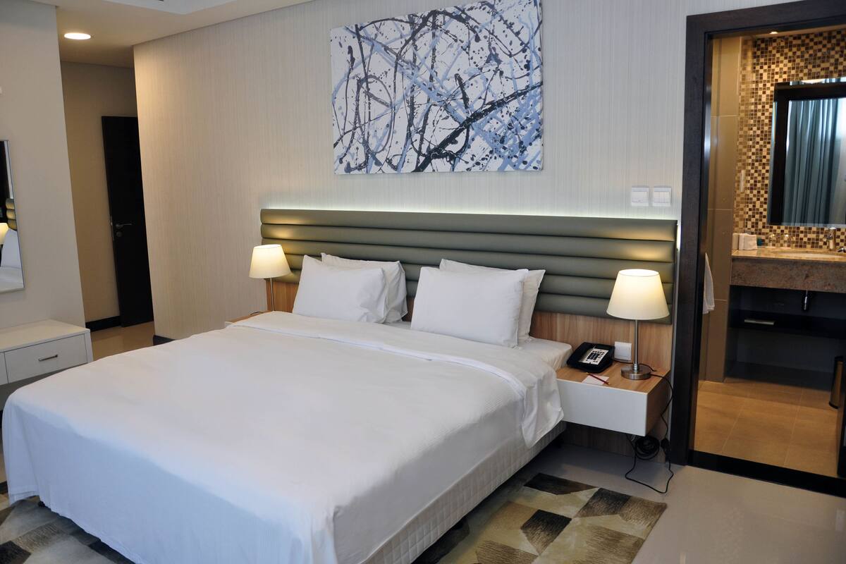 A hotel room with a large bed, two bedside lamps, a nightstand with a phone, a wall art piece, and a bathroom visible through an open door.