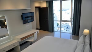 Una habitación de hotel con una cama grande, un escritorio con televisor de pantalla plana, un ventanal con vistas a edificios y una puerta que conduce a otra habitación.