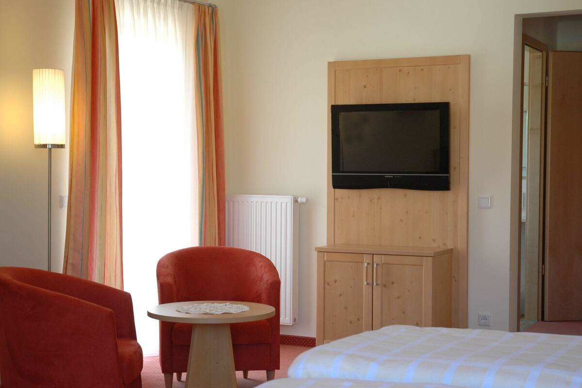 A hotel room with a bed, a television mounted on a wooden cabinet, a red armchair, a small round table, and a window with curtains.