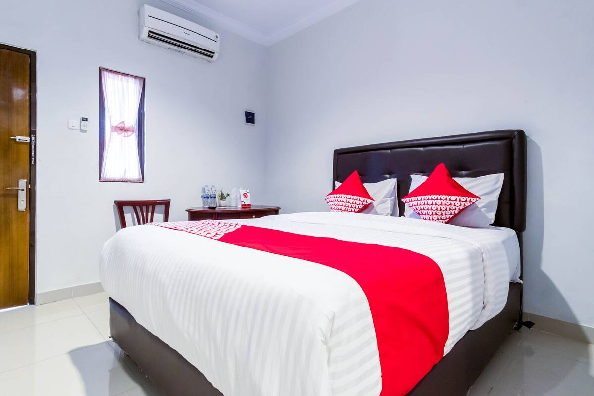A hotel room with a bed, a bedside table, a chair, a window with curtains, and an air conditioning unit.