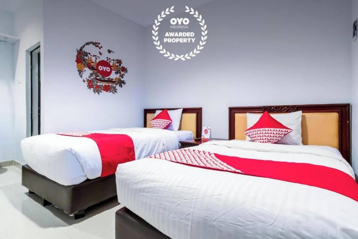 A hotel room with two beds, each with a red and white bedspread and matching pillows. A circular award plaque is visible on the wall.