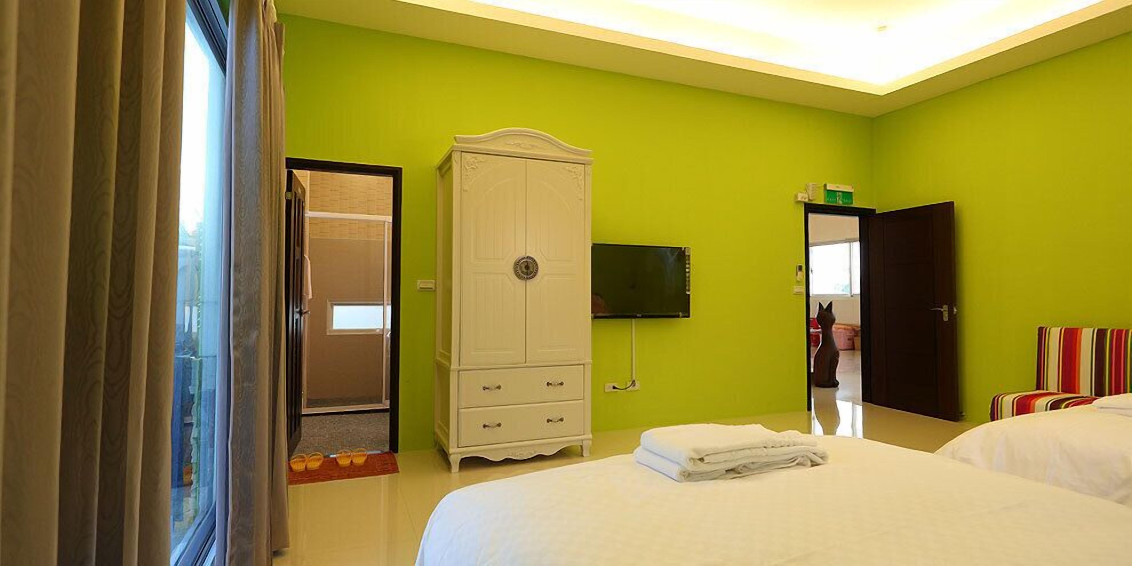 A hotel room with a green wall, a white wardrobe, a bed with white bedding, a TV mounted on the wall, and a door leading to another room.