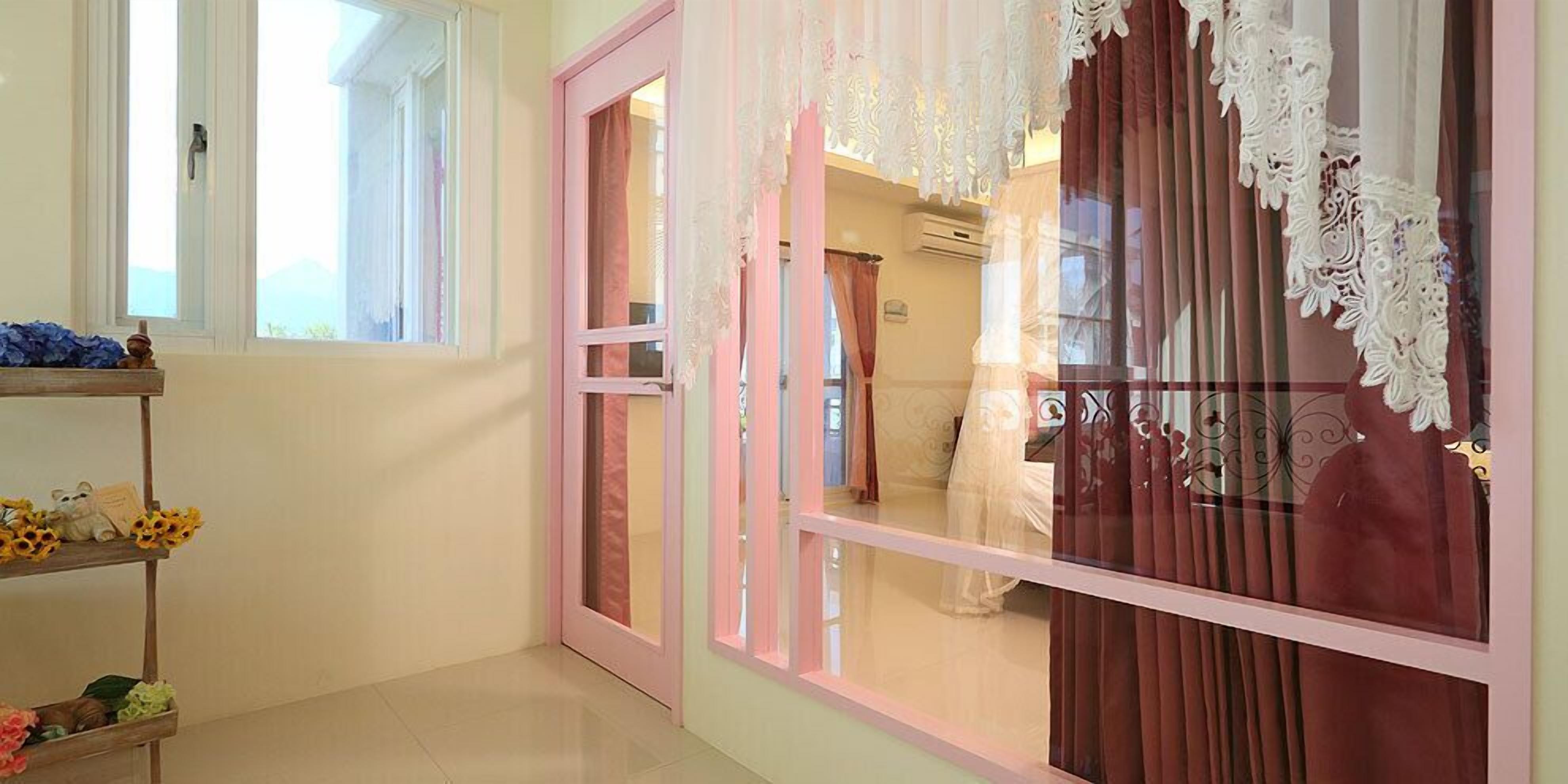 Una habitación con una ventana de marco rosa, una estantería de madera con objetos decorativos, y un vistazo a una cama con colcha blanca.
