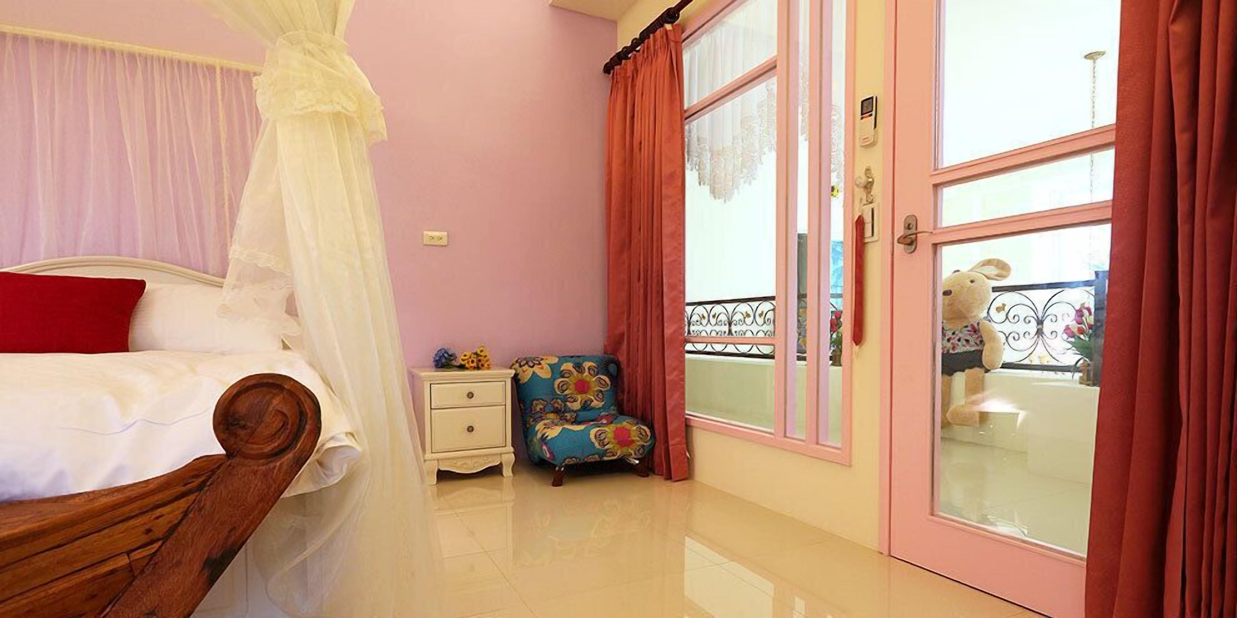 Un dormitorio con cama a dosel, una mesita de noche, una silla colorida y un ventanal con cortinas.