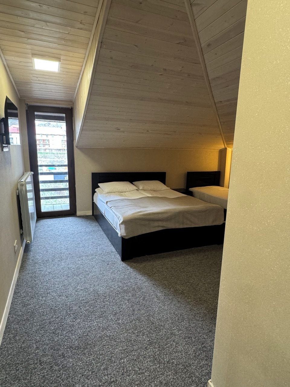 A small, single-room apartment with a sloped ceiling, a bed, a radiator, and a door leading to an outdoor area.