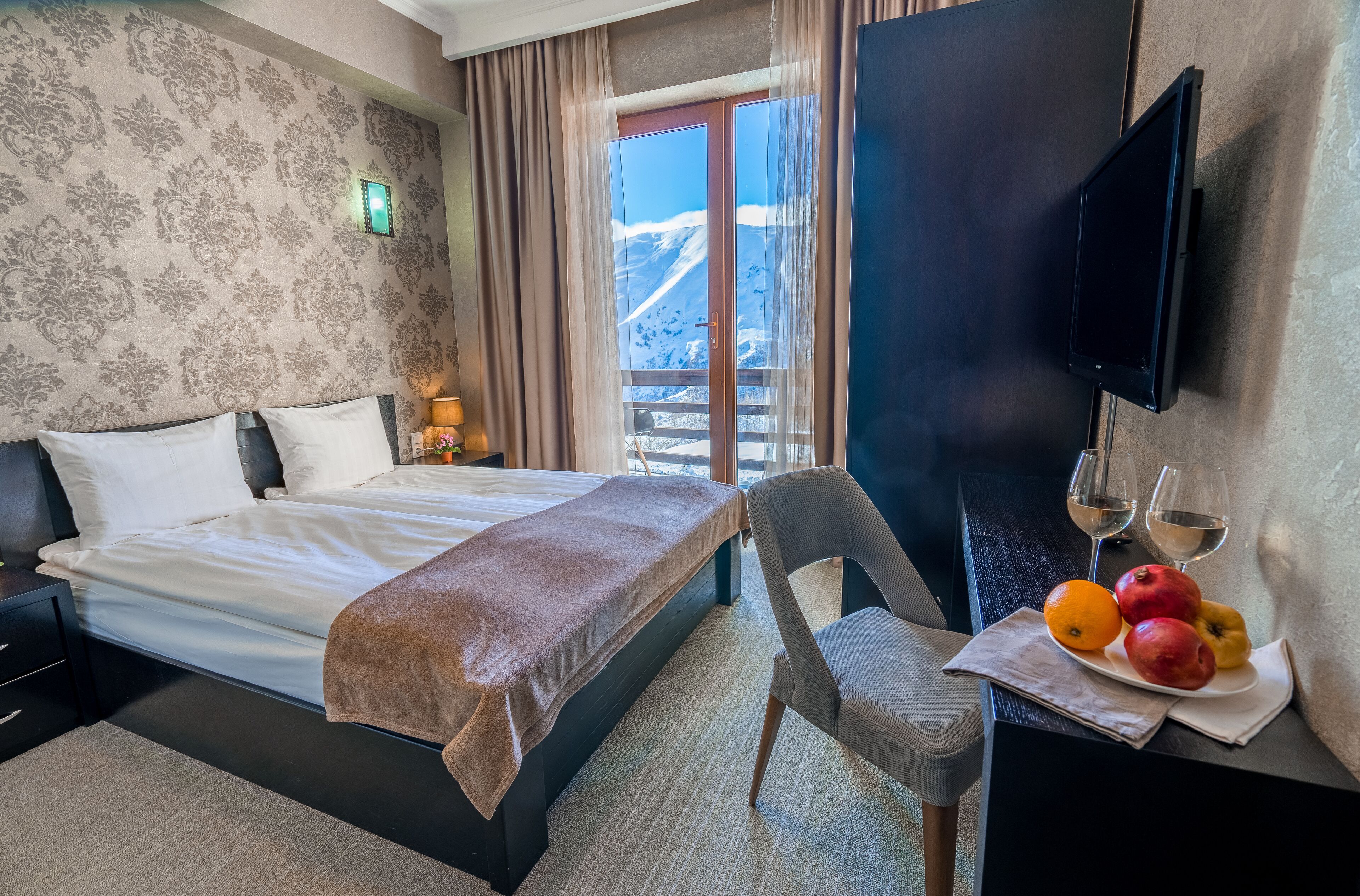 A hotel room with a bed, a chair, a dining table with fruit and wine glasses, and a TV on the wall.
