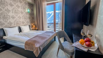 A hotel room with a bed, a chair, a dining table with fruit and wine glasses, and a TV on the wall.
