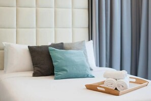 A neatly made bed with a wooden tray holding rolled towels, a tufted headboard, and a window with curtains.