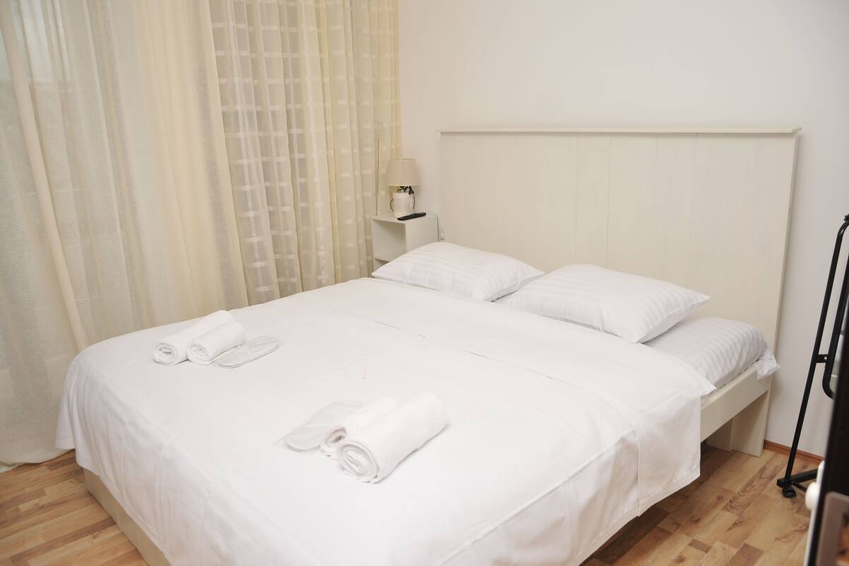 Una cama bien hecha con sábanas y toallas blancas, una mesita de noche con lámpara y un piso de madera.