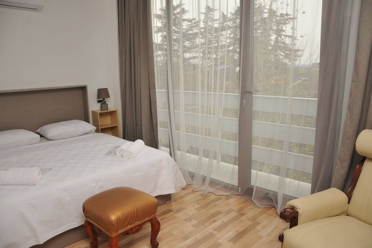Habitación de hotel con cama, mesita de noche de madera, una silla y un amplio ventanal con vistas a árboles.