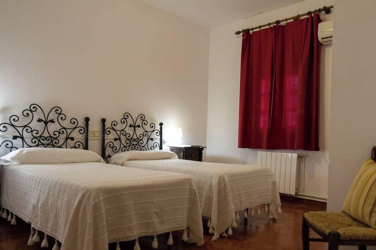 Quarto com duas camas, um radiador, uma janela com cortinas vermelhas e uma cadeira.