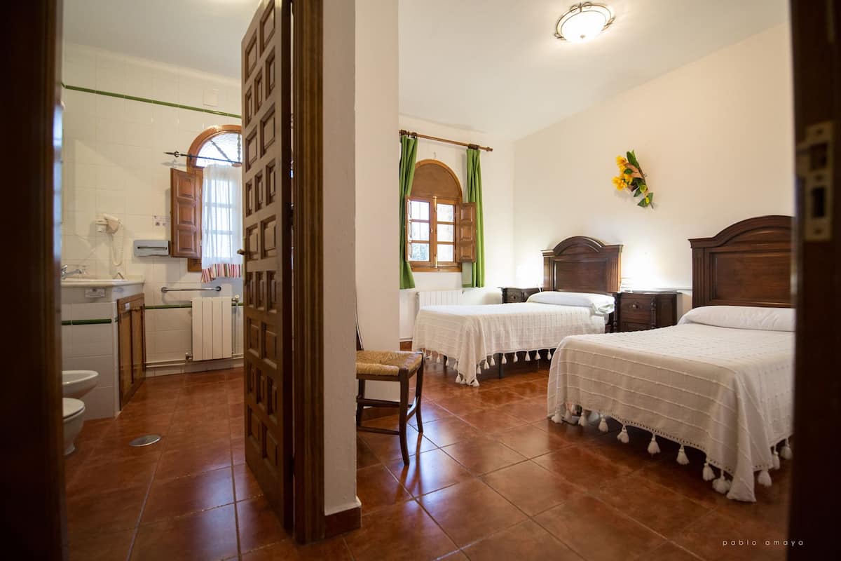 Quarto de hotel com duas camas, um espaço de estar pequeno, banheiro com vaso sanitário e pia, e janela com cortinas.
