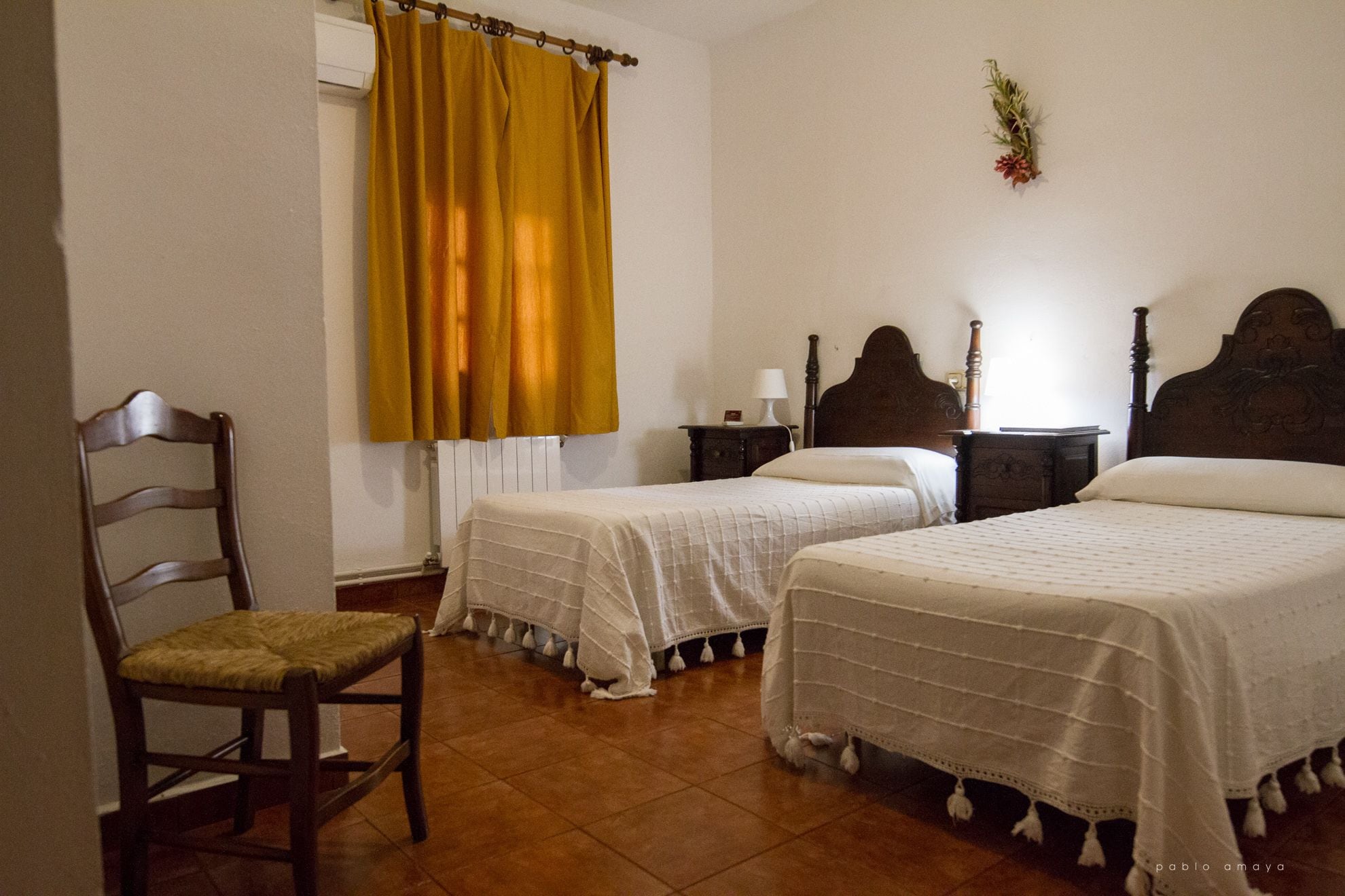 Un dormitorio con dos camas individuales, una silla de madera, una mesita de noche con una lámpara y una ventana con cortinas amarillas.