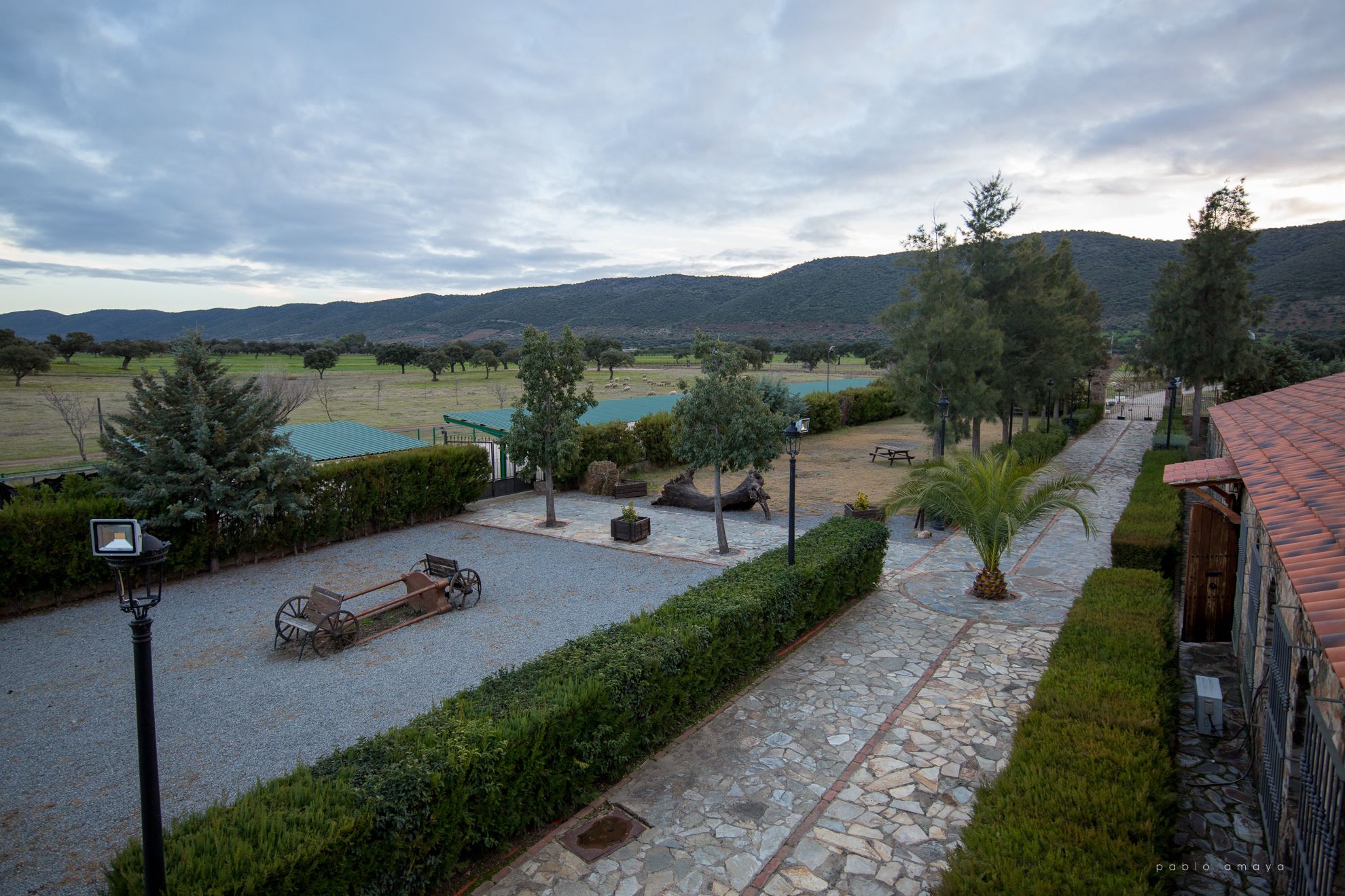 A courtyard with a stone pathway, a wooden cart, and a view of a lake and mountains.