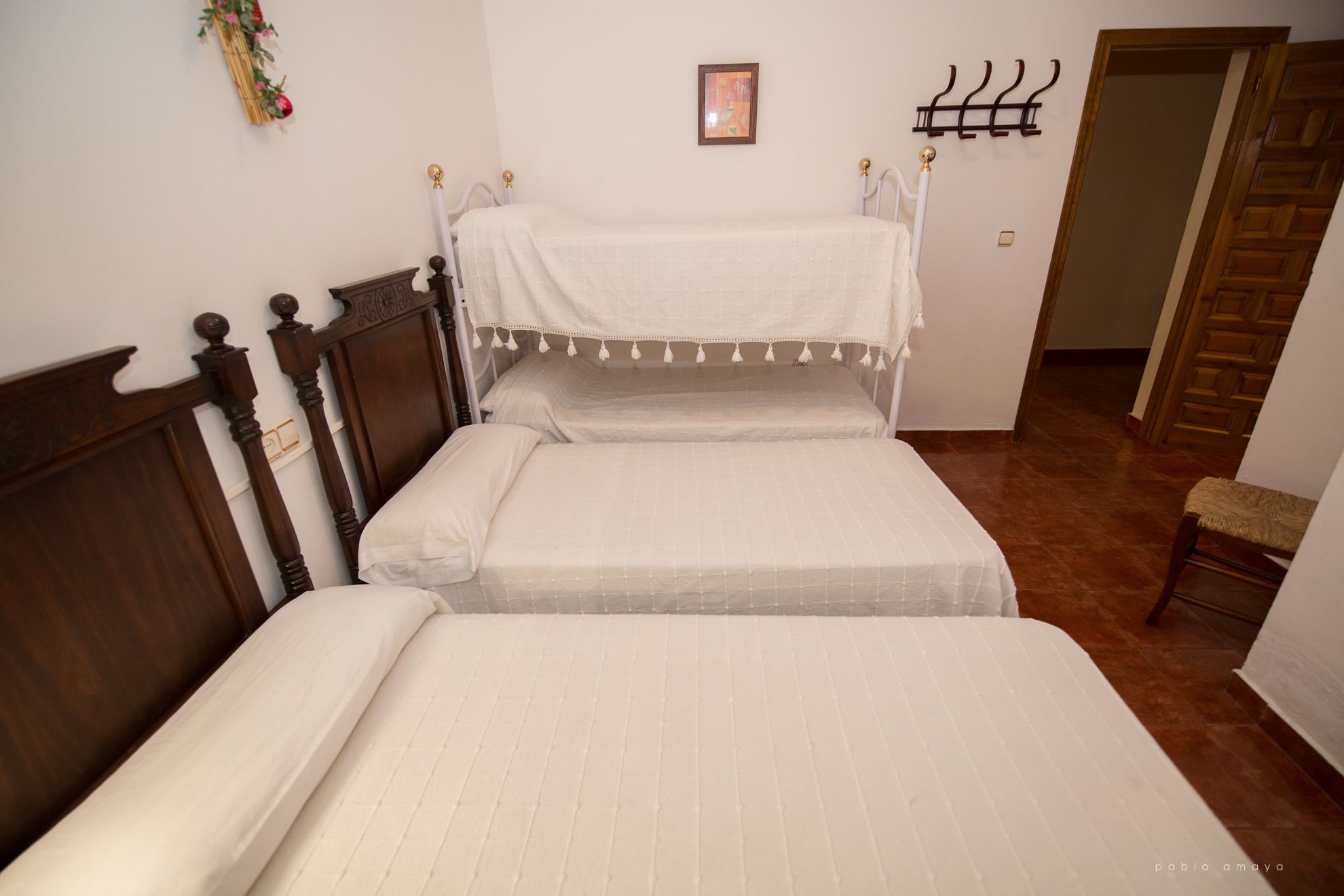 Un dormitorio con dos camas, cabeceros de madera, una mesita de noche pequeña, un perchero en la pared y un cuadro enmarcado.