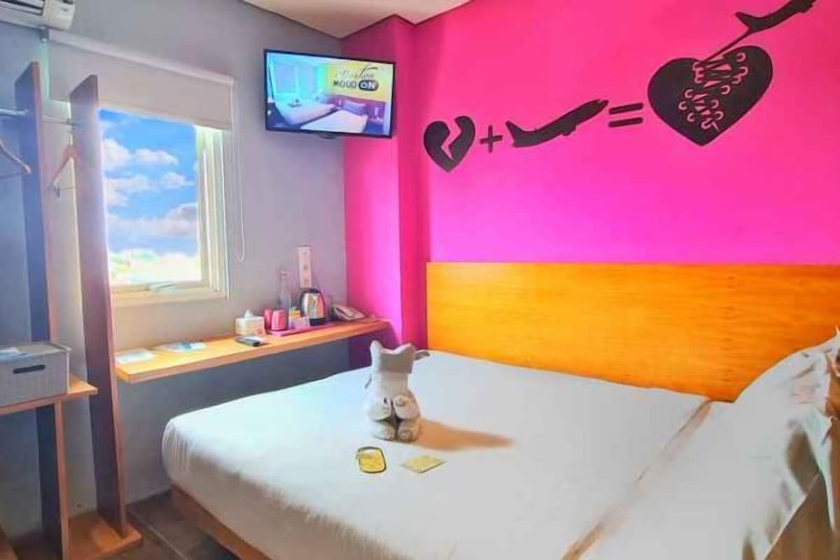 A bedroom with a bed, a desk with a computer, a TV, and a wall decoration.