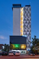 Egy modern hotel, amelynek geometrikus homlokzata van, jól látható étterme, valamint egy parkoló, ahol több autó is elfér.
