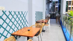 En overdækket udendørs siddeplads med et langt bord og fletstole, en mønstret væg og en glasgelænder med udsigt over en have.