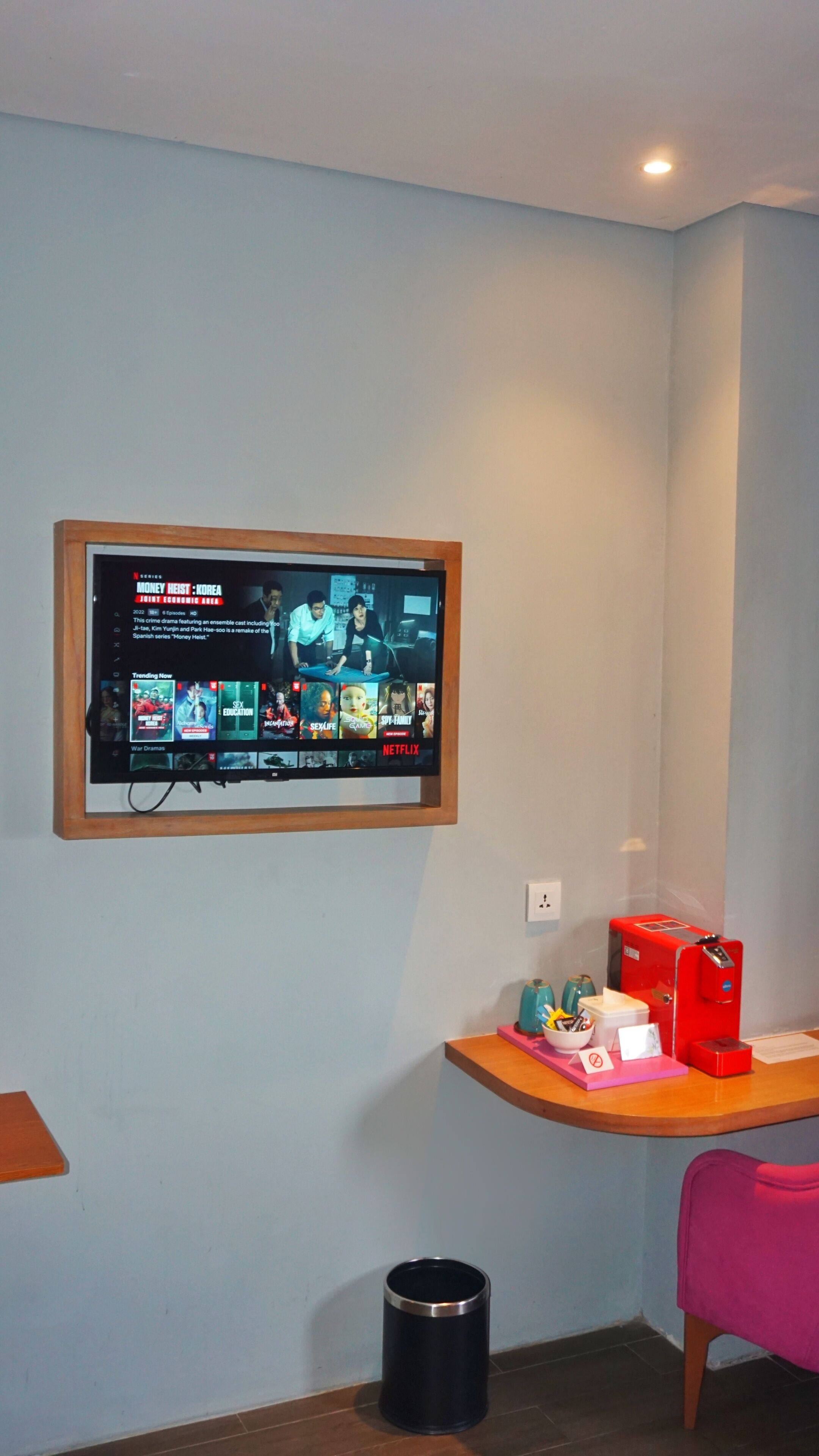 Quarto com televisão fixada na parede exibindo a interface de um serviço de streaming, uma escrivaninha de madeira com um objeto vermelho, uma lixeira e uma cadeira rosa.