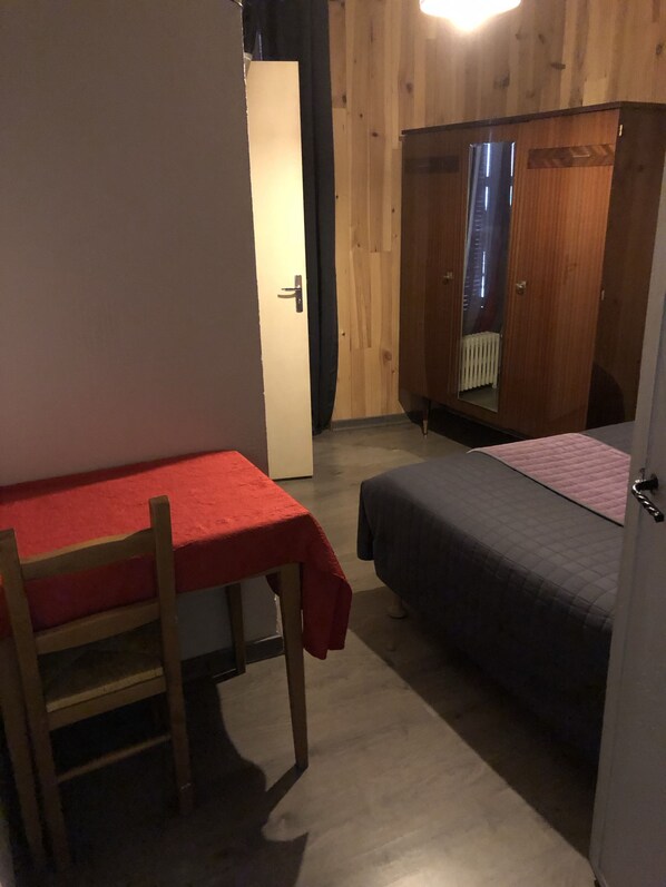 Una habitación pequeña con un armario de madera, una cama, una mesa con mantel rojo y una silla.