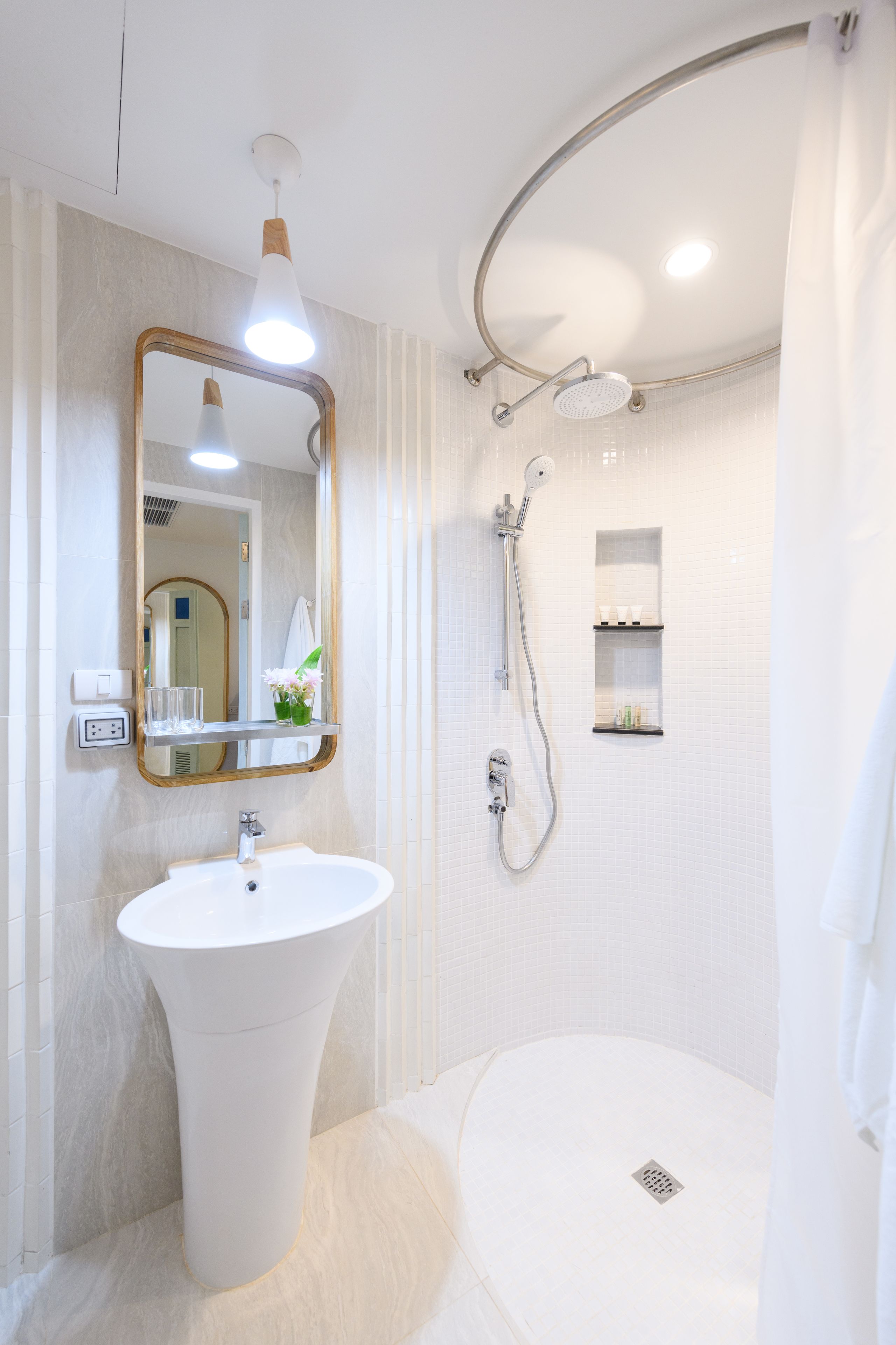 A modern bathroom with a pedestal sink, a large mirror, a shower with a handheld showerhead, and built-in shelves.