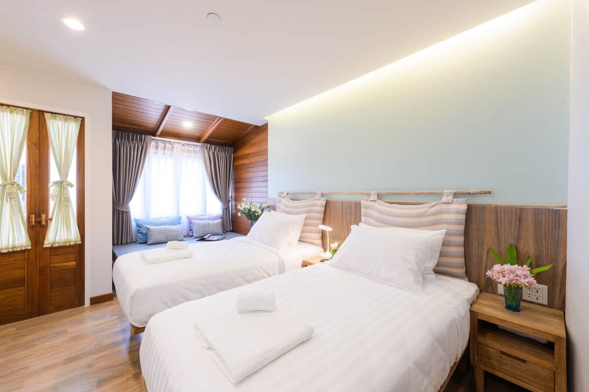 Une chambre d’hôtel avec deux lits, des têtes de lit en bois, une table de chevet avec une lampe et une fenêtre avec des rideaux.