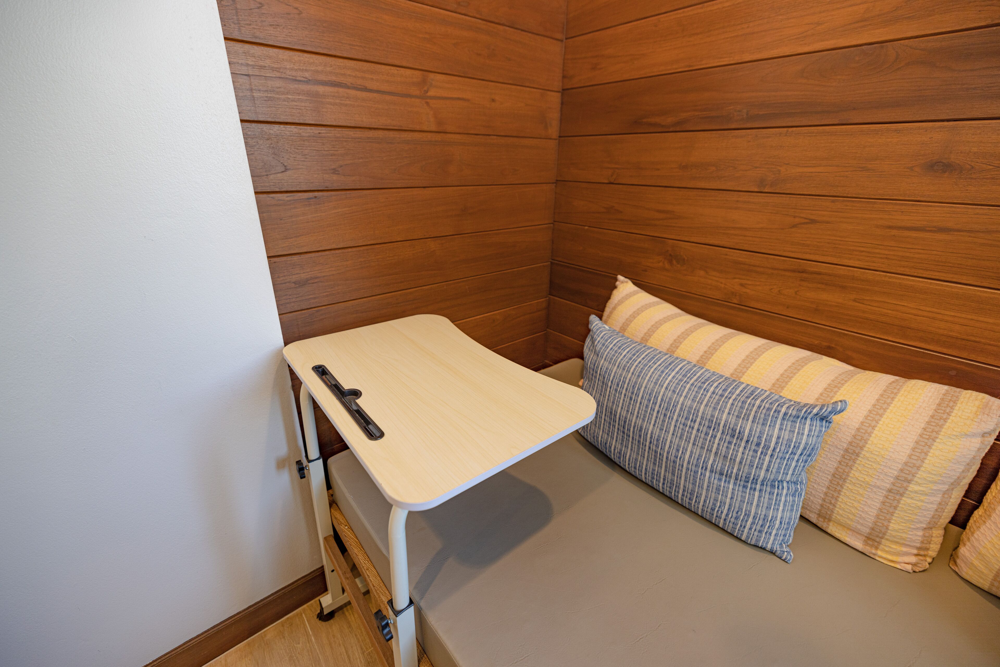 A corner room with a wooden wall, a small bed with striped pillows, and a white table with a black object on it.