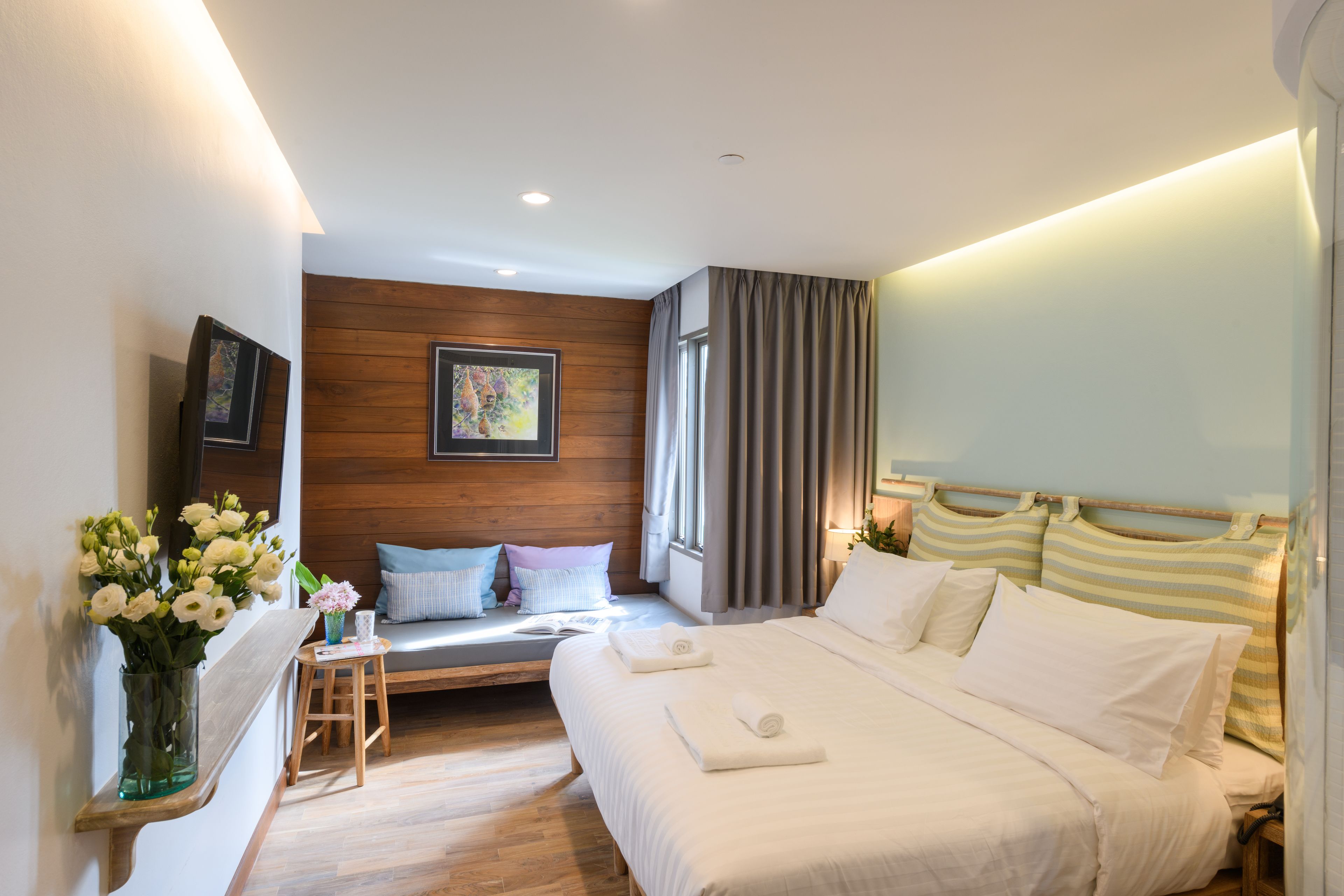 Ein modernes Hotelzimmer mit einer Holzvertäfelung, einem Bett mit weißer Bettwäsche, einem Nachttisch mit Blumen in einer Vase und einem Fenster mit Vorhängen.