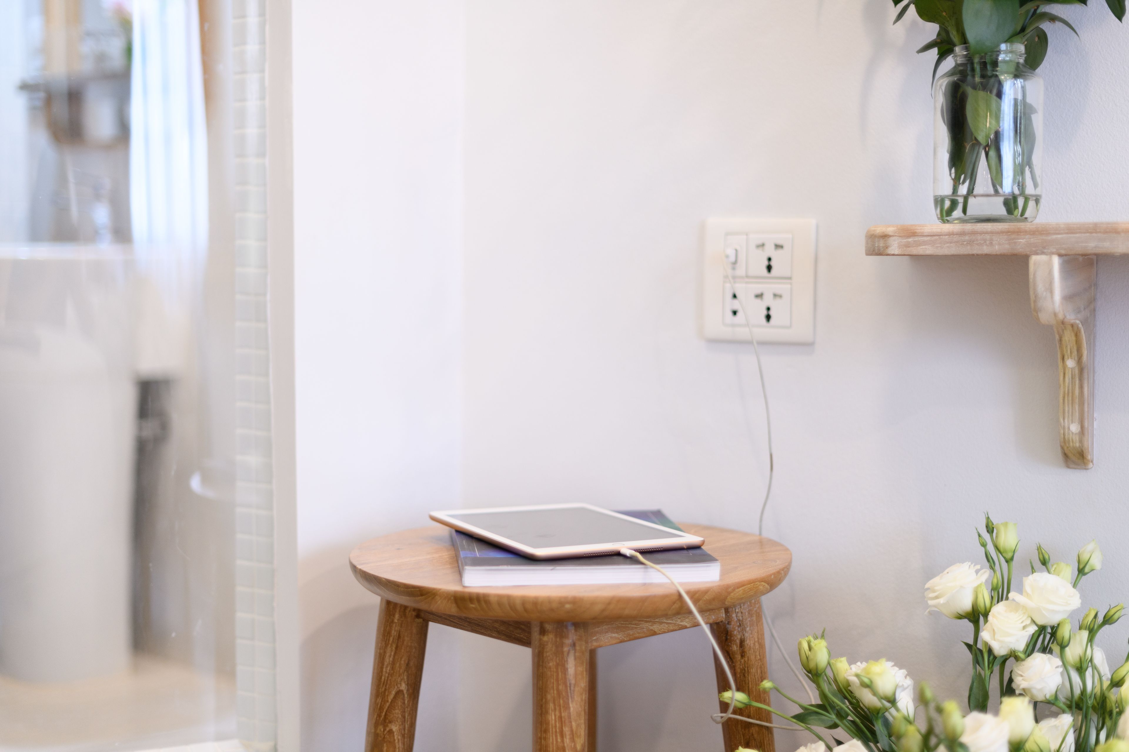 Ein Holzhocker mit einem Tablet und Ladegerät, eine Vase mit weißen Blumen und eine Steckdose an einer weißen Wand.