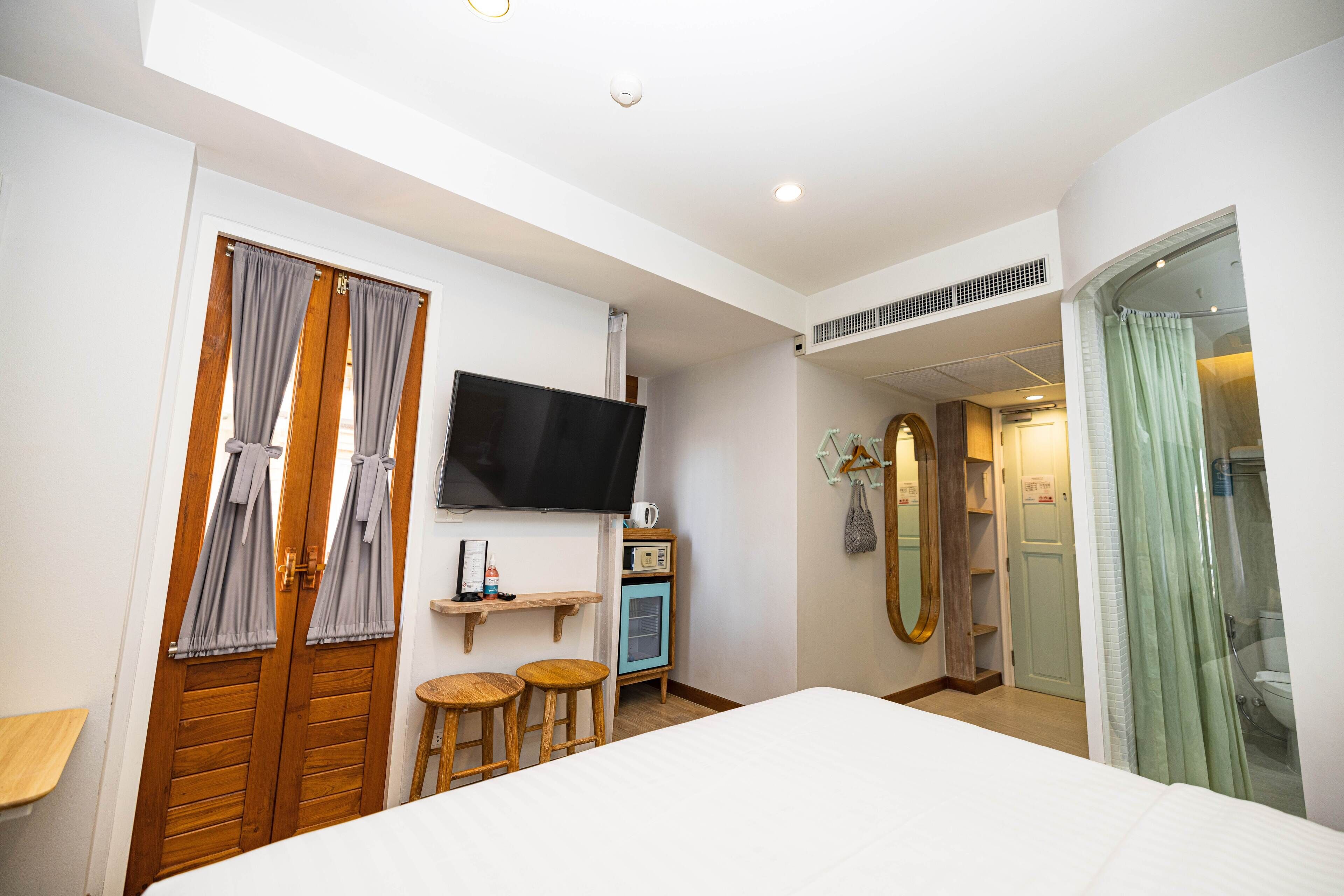 A bedroom with a bed, a TV, a small table, two stools, a shelf with a radio, and a mirror.