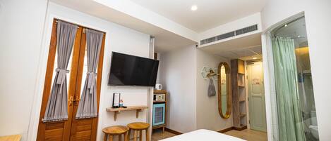 A bedroom with a bed, a TV, a small table, two stools, a shelf with a radio, and a mirror.