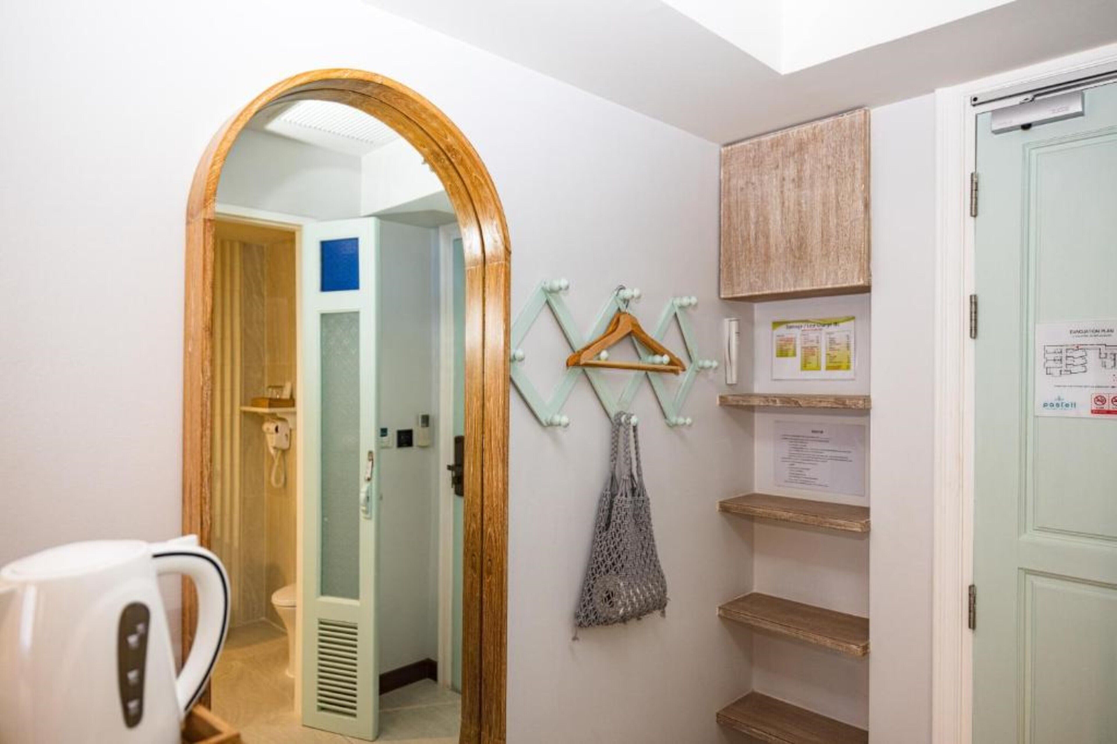 Quarto de hotel com porta de madeira, espelho, cabideiro com ganchos, chaleira e uma pequena estante com papéis.