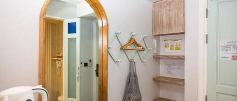 Quarto de hotel com porta de madeira, espelho, cabideiro com ganchos, chaleira e uma pequena estante com papéis.