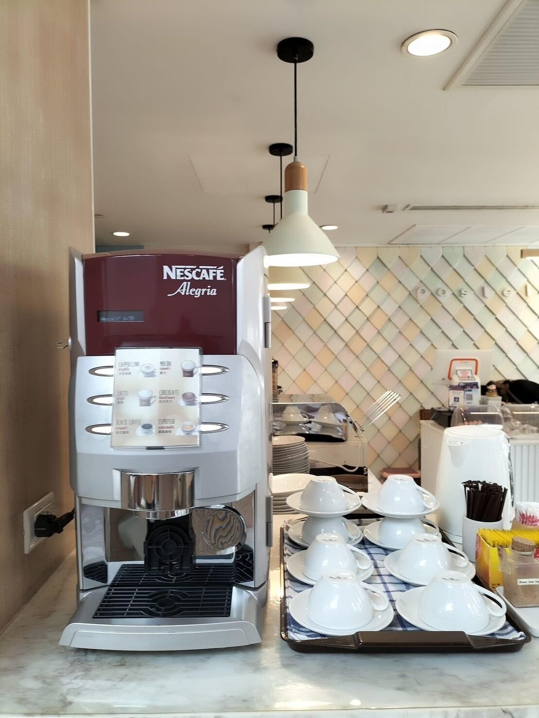 A coffee machine with multiple drink options, a tray of white cups, and a wall with a diamond pattern.