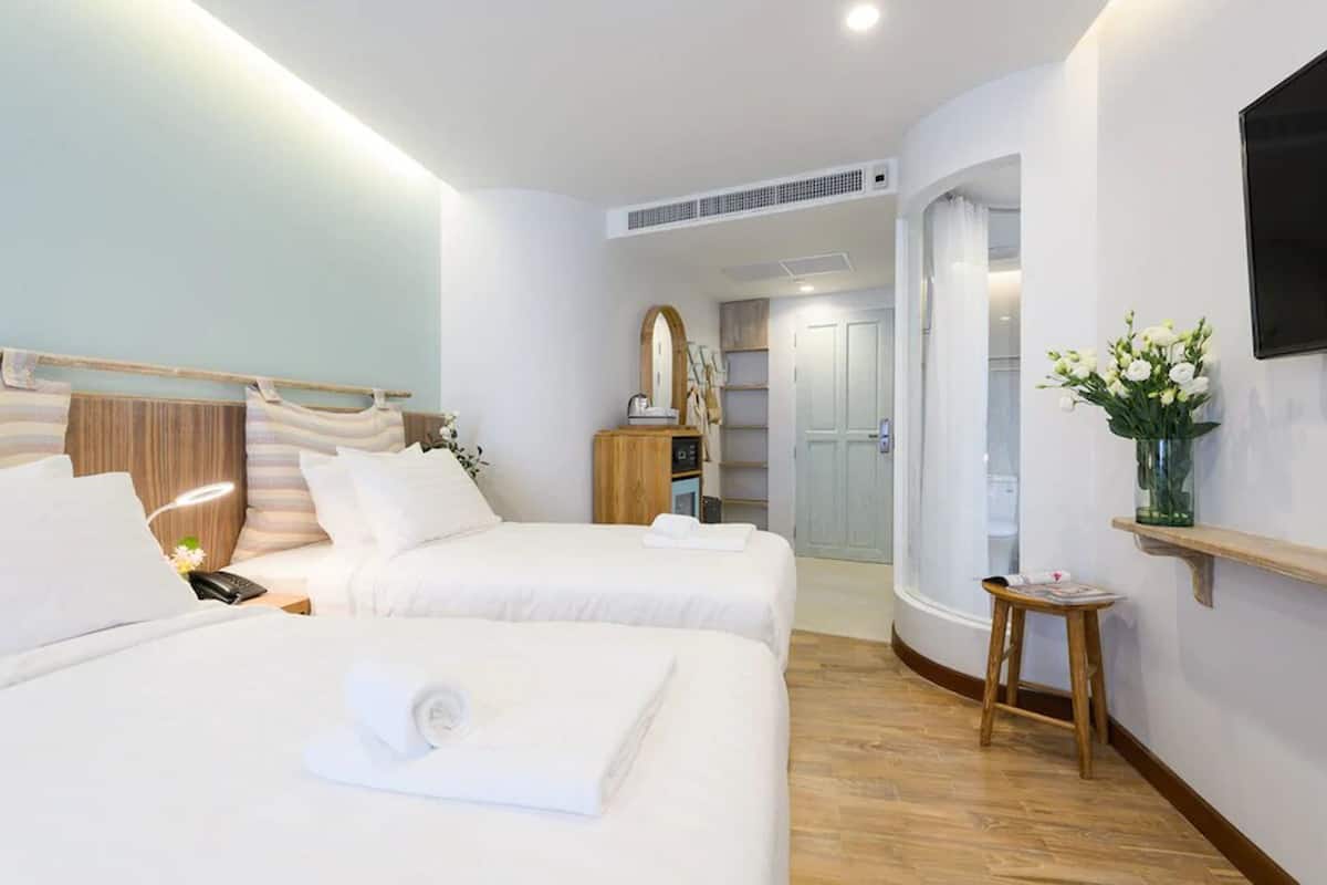 Une chambre d’hôtel avec deux lits, une tête de lit en bois, une table de chevet avec une lampe, un miroir, un petit tabouret et un téléviseur fixé au mur.