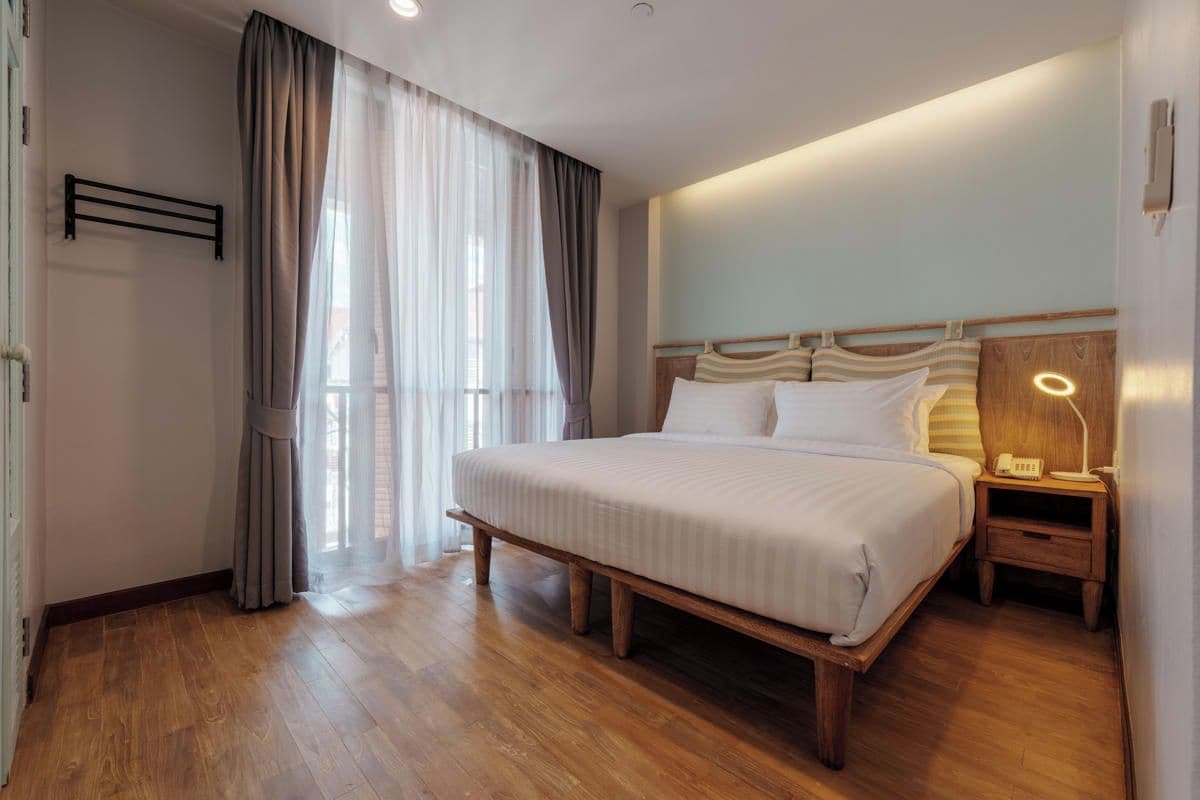 A hotel room with a large bed, wooden headboard, a bedside table with a lamp, and a window with curtains.