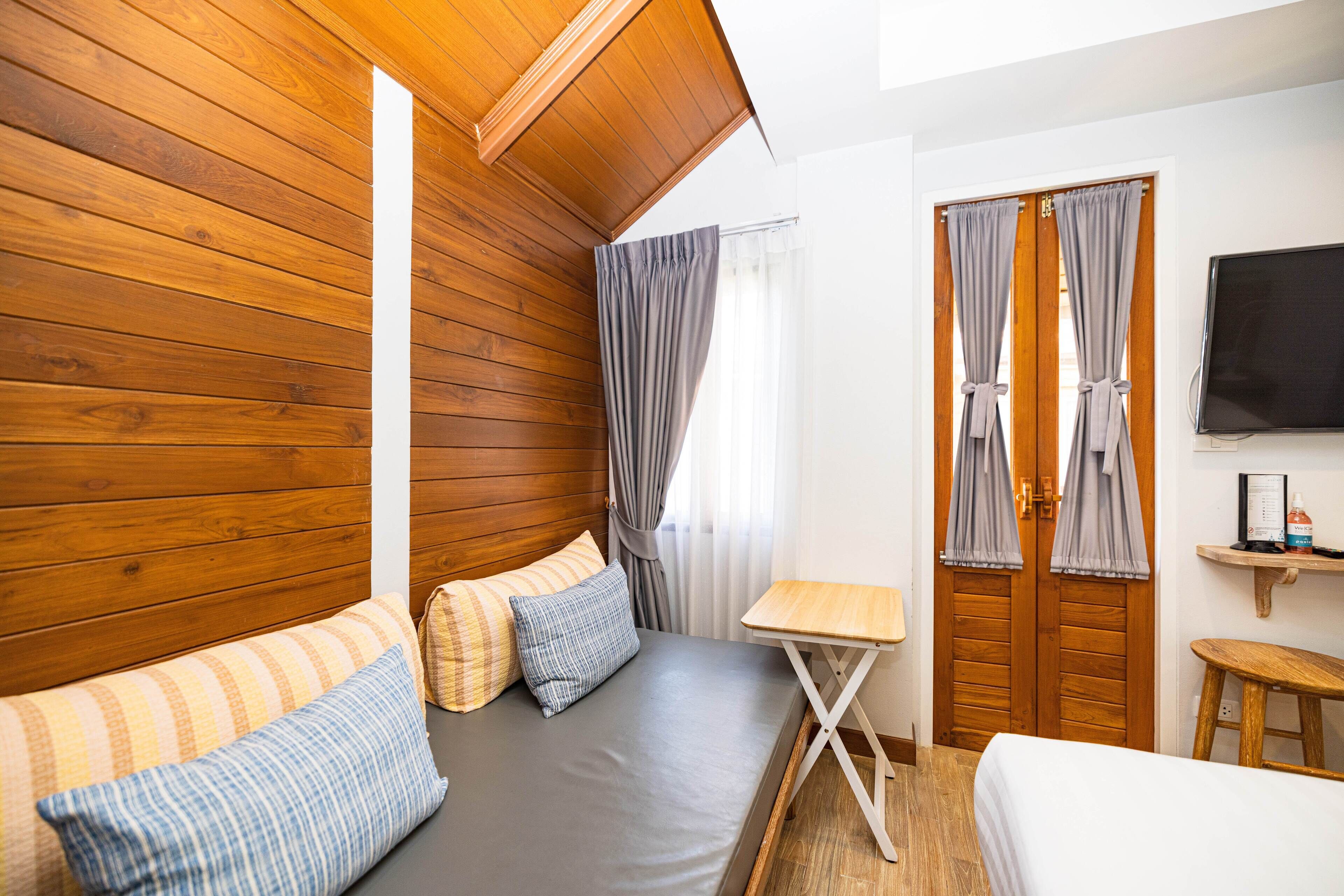 A room with a wooden ceiling, a bed with striped bedding, a small table, and a TV on the wall.