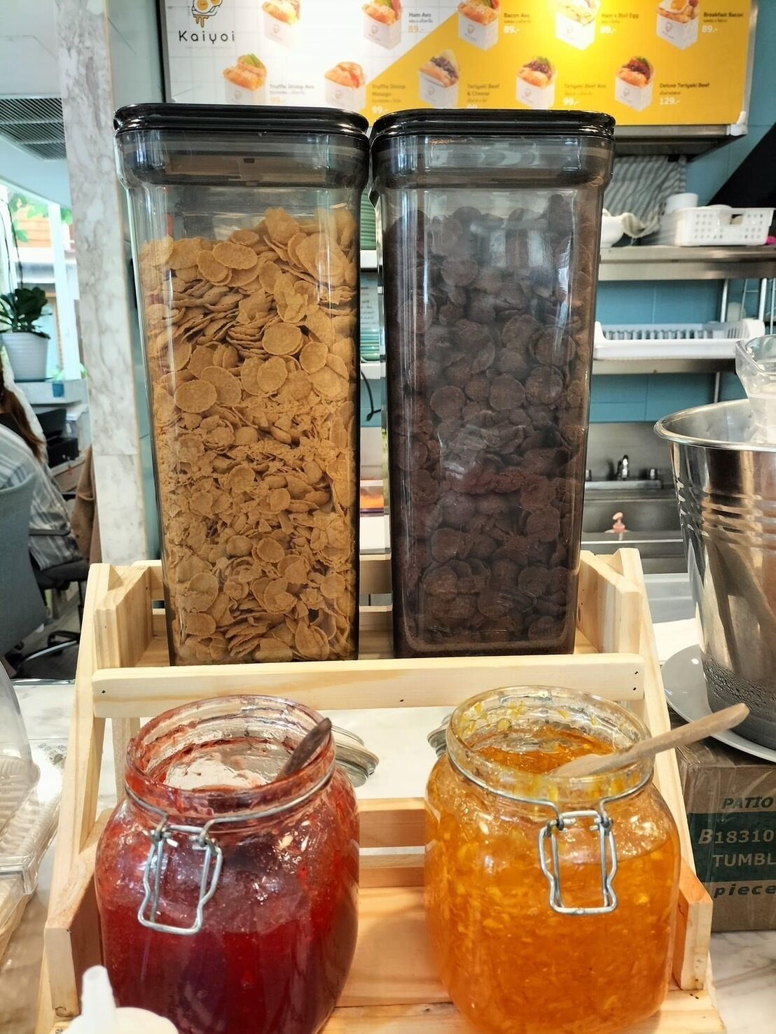 A food display with cereal and cookies in clear containers, and jars of red and orange preserves on a wooden stand.