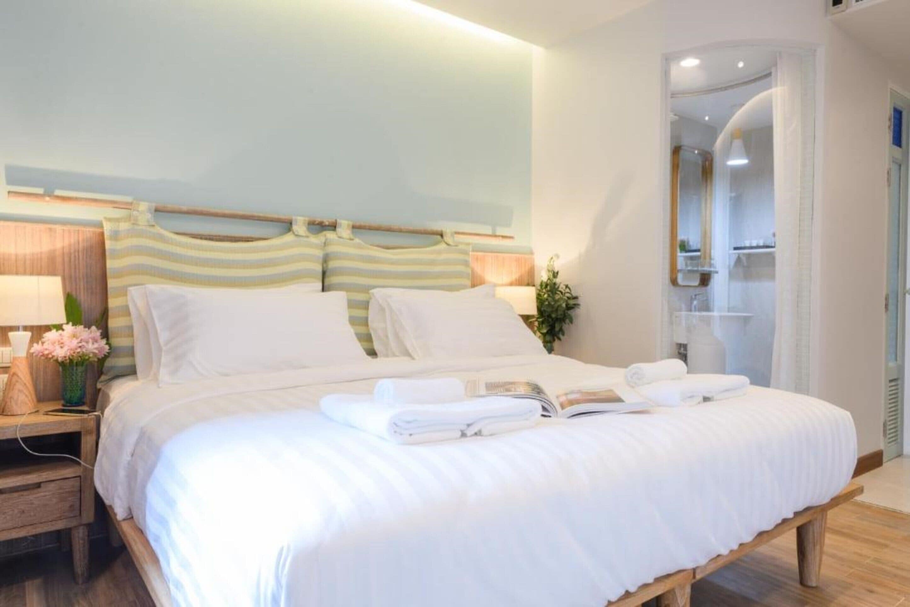 Una cama bien hecha con sábanas blancas, una mesita de noche con lámpara y un jarrón de flores, y un espejo grande en la pared.