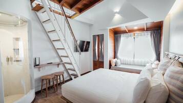 Un dormitorio con una cama, una ventana con cortinas, un televisor, una escalera de madera y una pequeña zona de estar con taburetes.