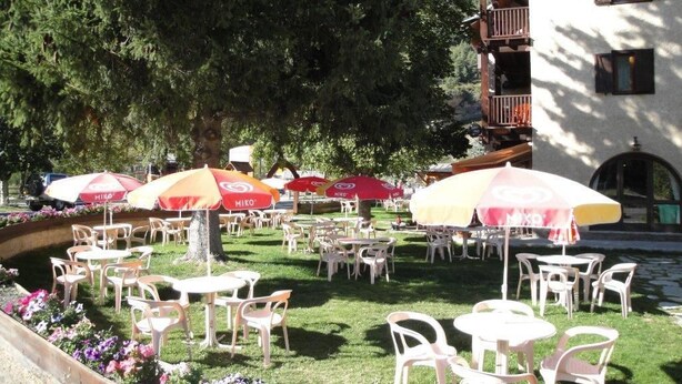 An outdoor seating area with white plastic chairs and tables, surrounded by flower beds and umbrellas. - Hotel Le Guilazur (Chateau-Ville-Vieille)