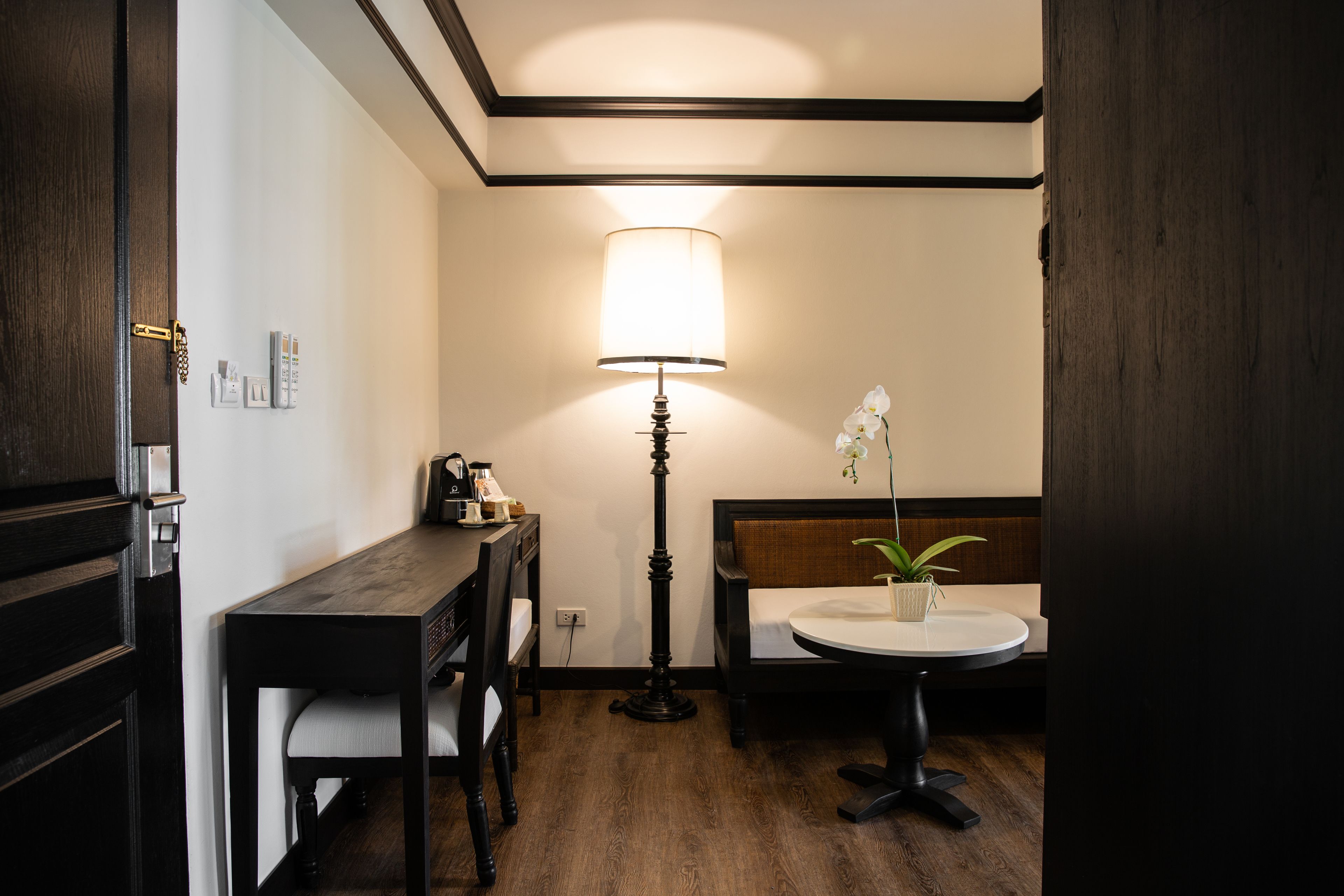 Une pièce comprenant un bureau en bois, une chaise, une table de chevet avec une lampe, une petite table ronde et un lit avec une tête de lit.