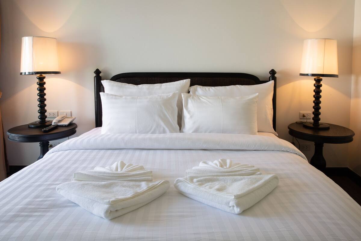 Una cama bien hecha con sábanas blancas, dos lámparas de noche y toallas dobladas sobre la cama.