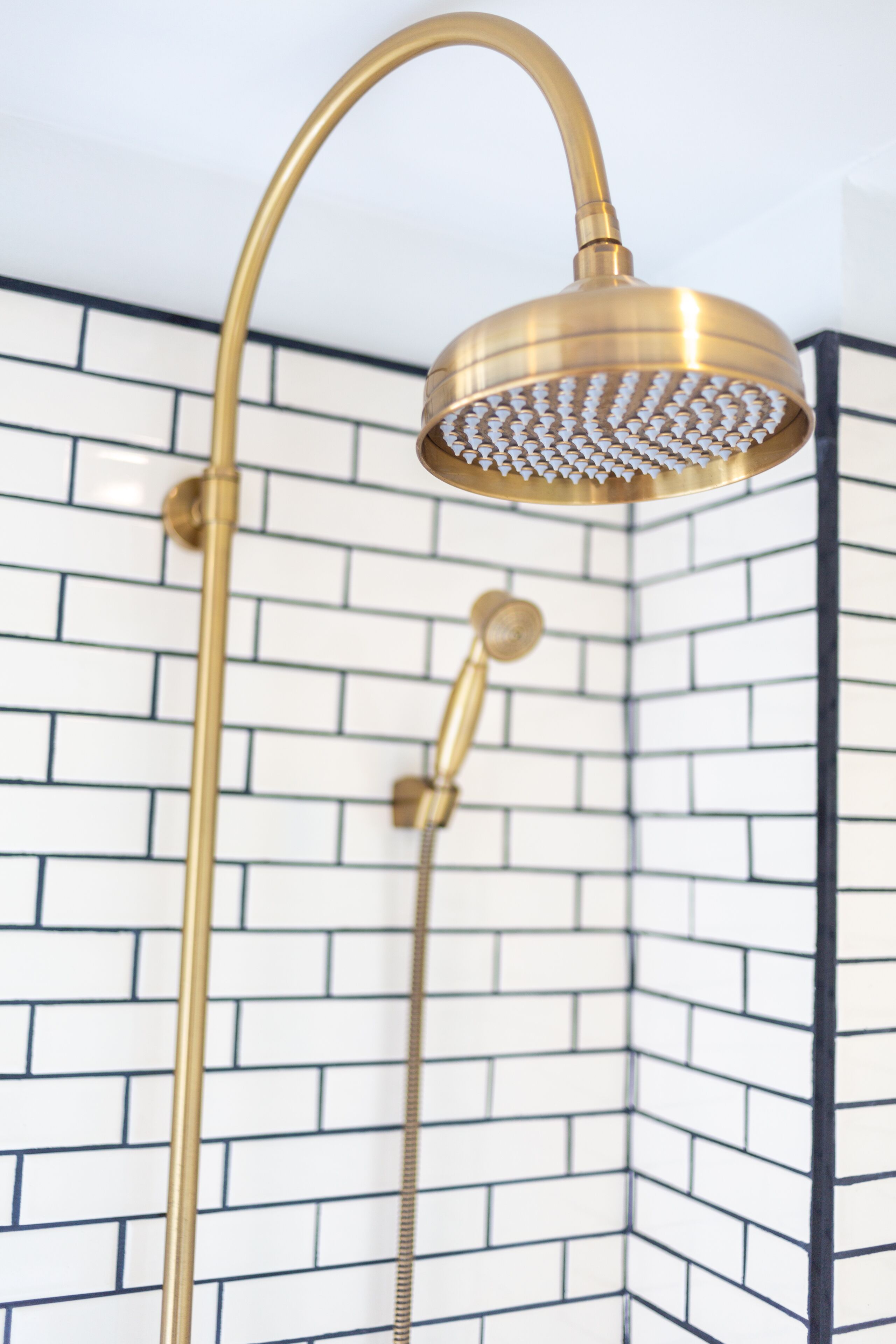 Esquina de un baño con ducha de oro y cabezal de ducha de mano, frente a una pared de azulejos blancos con lechada negra.