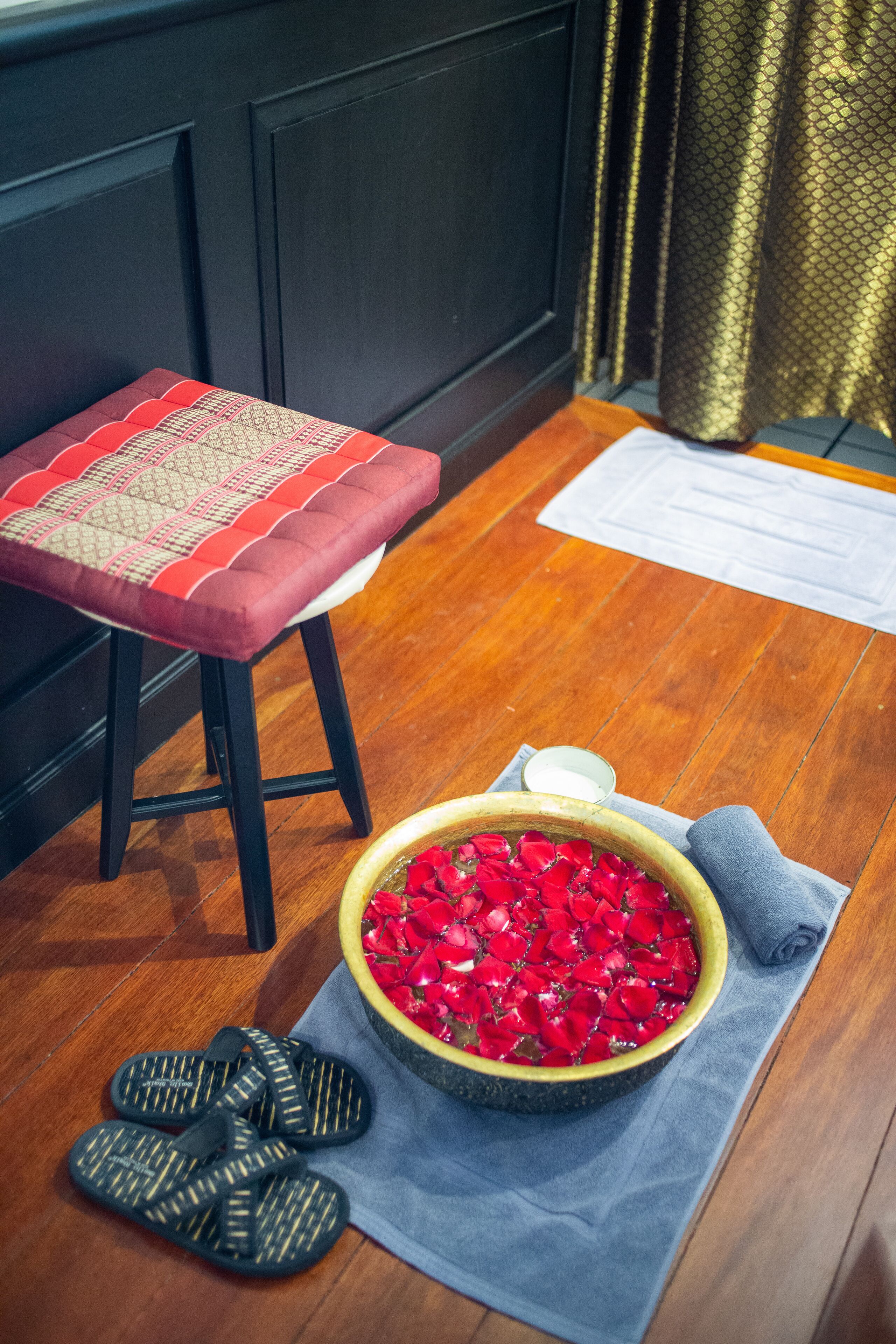 A wooden floor, a red cushioned stool, a bowl of red rose petals, a small bowl, and black sandals.