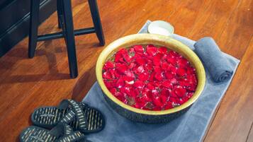 A wooden floor, a red cushioned stool, a bowl of red rose petals, a small bowl, and black sandals.