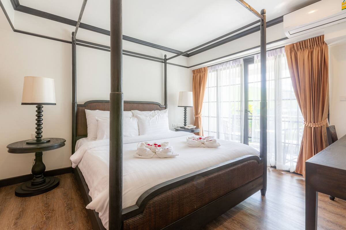 Una cama con dosel, ropa de cama blanca, dos mesitas de noche con lámparas y vistas al exterior a través de grandes ventanales.